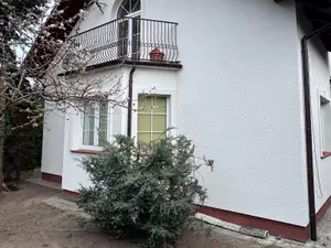 Ściana domu jednorodzinnego z białym tynkiem cienkowarstwowym, kutą balustradą balkonu i ceglaną podmurówką