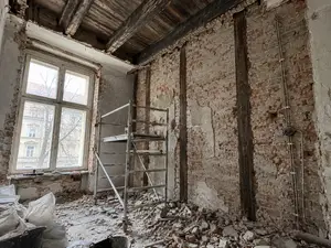 Wnętrze mieszkania w kamienicy w trakcie remontu z odsłoniętym drewnianym stropem belkowym, skutymi tynkami i starymi przewodami na ścianie