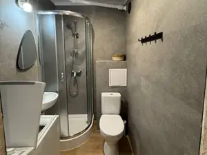 Mała łazienka po remoncie z półokrągłą kabiną prysznicową, umywalką, WC kompaktowym i obudową z białą klapą rewizyjną, ściany w szarych płytkach