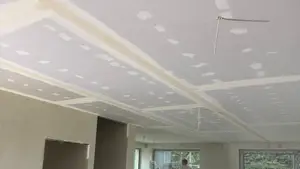 Sufit z płyt gipsowo-kartonowych w trakcie szpachlowania spoin i łbów wkrętów, z wyprowadzonymi przewodami do lamp, w jasnym wnętrzu domu z dużymi oknami